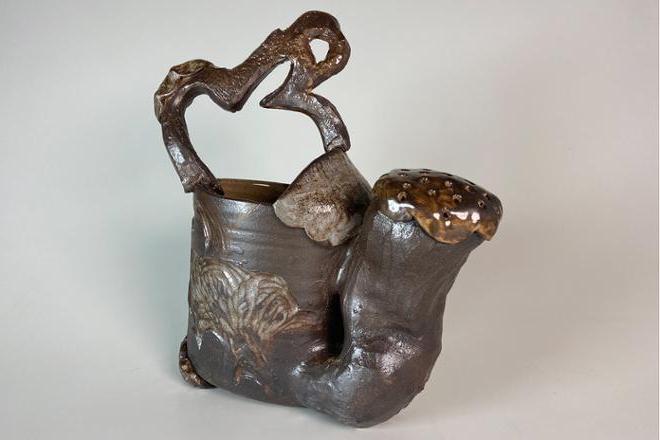 Dark ceramic watering can with loopy handle and complimentary carvings surrounding the base of the from. These carvings are brought out by a lighter hued shino glaze.